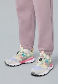 Fötter i pastellfärgade sneakers med grå, rosa och blå detaljer, tillsammans med ljuslila träningsoverallsbyxor och vita strumpor på ett grått golv.