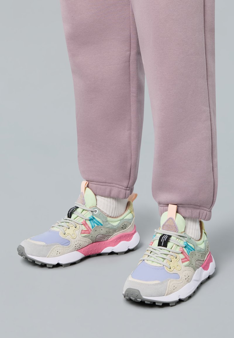 Fötter i pastellfärgade sneakers med grå, rosa och blå detaljer, tillsammans med ljuslila träningsoverallsbyxor och vita strumpor på ett grått golv.