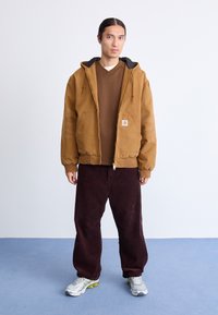 Une jeune personne avec de longs cheveux, portant une veste à capuche marron, un shirt marron, un pantalon large foncé et des baskets blanches, se tient sur un sol bleu contre un mur blanc.