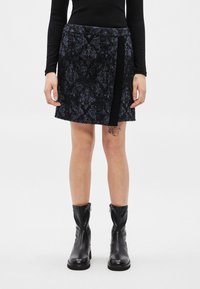 Femme portant un haut noir à manches longues, une jupe noire et bleue à motif, des bottes en cuir noires jusqu'au milieu du mollet et des tatouages visibles sur les jambes.