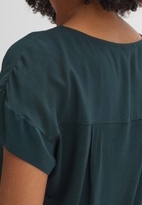 Tmavě zelená halenka s volným střihem, krátkými ohrnutými rukávy a hladkou, plynulou texturou s jemným leskem.