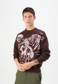 Suéter de punto marrón con un diseño gráfico rosa de un jinete contra un fondo montañoso; cuello y puños acanalados.