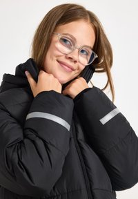 Fiatal nő áttetsző szemüvegben, fekete steppelt kabátot viselve, a gallért közel tartva magához, fehér háttér előtt.