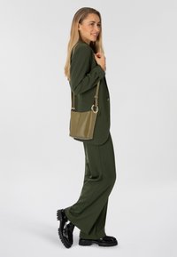 Donna in completo verde oliva e scarpe nere, che porta una borsa a tracolla verde oliva, in piedi di lato e sorridente su sfondo bianco.