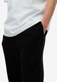 Schwarze Sweatpants mit einer lockeren Passform, ausgestattet mit seitlichen Taschen und einem dezenten Logo auf der linken Seite. Kombiniert mit einem weißen T-Shirt.