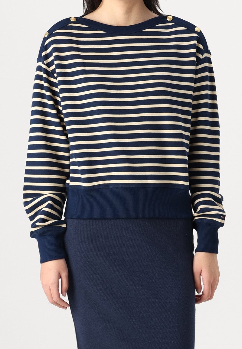 Femme portant un haut à manches longues rayé bleu marine et beige avec des boutons dorés sur les épaules et une jupe bleu marine.