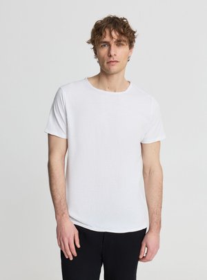 Giovane uomo con capelli ricci che indossa una t-shirt bianca semplice a maniche corte e pantaloni neri, in piedi davanti a uno sfondo grigio chiaro.