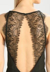 Robe sans manches en dentelle noire avec des motifs floraux complexes et un dos ouvert prononcé, portée par une personne à la peau claire et aux cheveux blonds.