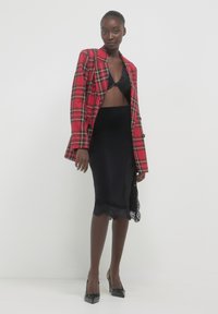 Rot karierter Blazer, schwarzer Rock mit Spitzenbesatz, schwarzes Bralette und spitze schwarze High Heels. Glattes Material, strukturiertes Design, geschichtete Texturen.