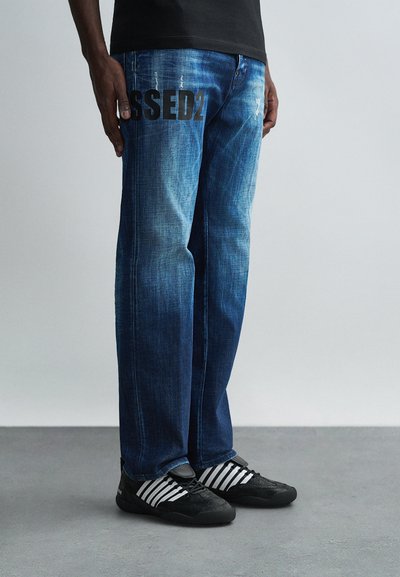 Jeans in denim blu con texture sbiadita, taglio dritto e una grande grafica nera "BLESSED" stampata sulla gamba. Scarpe sportive nere con dettagli a strisce.