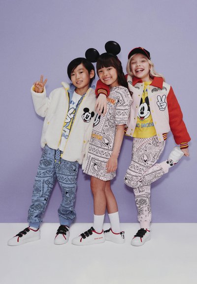 Drie kinderen dragen kleurrijke outfits met ontwerpen van Mickey Mouse. De stoffen omvatten pluizig wit, gestreepte sweatpants en bijpassende schoenen.