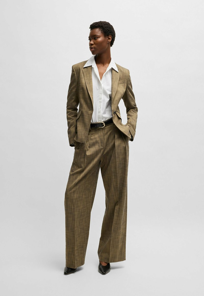 Traje a cuadros en tono beige con corte entallado, que presenta una camisa blanca de contraste debajo y un cinturón negro. Modelo llevando tacones negros puntiagudos.