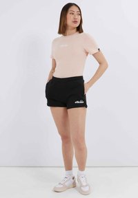 Camiseta ajustada de color rosa claro con logo blanco, combinada con pantalones cortos deportivos negros con bolsillos laterales y logo blanco, completada con zapatillas casuales.