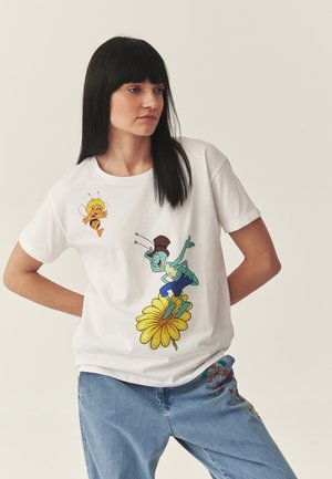 Kobieta o czarnych włosach ubrana w białą koszulkę z kreskówkową pszczołą i konikiem polnym na żółtym kwiatku, do tego niebieskie dżinsy z kwiatową haftem.