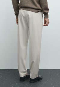 Light beige trousers with a relaxed fit, featuring a smooth texture and a clean design. Worn with black loafers, viewed from the back.