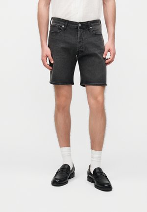 Mand iført sorte cowboyshorts, hvide sokker, sorte loafers og en hvid skjorte, stående mod en ensfarvet hvid baggrund.