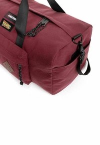 Borsa a duffel color marrone con forma rettangolare, superficie in tessuto, chiusura a doppia zip, manico nero e tasca laterale con un patch decorativo.