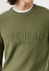 Alyvuogių spalvos megztinis su apvalia kaklo iškirpte ir tekstūruotu logotipu "PEPE JEANS LONDON" tamsesnėje atspalvyje, su gaubtu ir apykakle su raibais. 