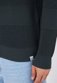 Pull en tricot texturé vert foncé avec des manches et un ourlet côtelés. Présente des motifs à rayures horizontales. Petite étiquette de logo visible au niveau de la taille.
