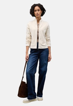 Hellbeige Reißverschluss-Strickjacke über einem sandfarbenen Top, kombiniert mit dunkelblauen weiten Jeans, cremefarbenen Sneakern und einer braunen Umhängetasche.
