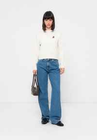 Pulover alb cu mâneci lungi, cu dungi, având o mică broderie cu un ursuleț brun, asortat cu blugi largi de culoare albastră deschisă și o geantă mică de umăr de culoare închisă.