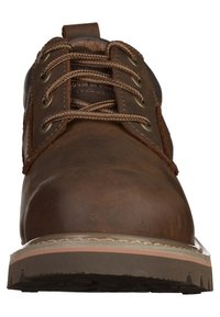 Scarpa in pelle marrone con punta arrotondata, lacci circolari, occhielli metallici e suola intermedia bianca. Parte superiore testurizzata con accenti cuciti.