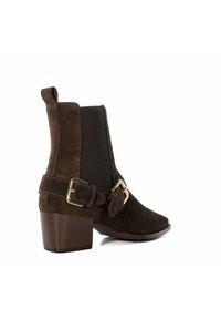 Bottine en suède marron avec un talon épais, des panneaux latéraux élastiques et une sangle décorative avec boucle. Le design est élégant et moderne.