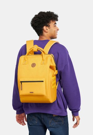 Sac à dos jaune avec un design carré, poignée supérieure, sangles latérales et poche zippée à l'avant. Fabriqué en tissu durable avec une texture lisse.