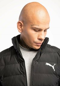 Czarna pikowana kurtka z stójką, z białym logo Pumy. Nosi się na szarym T-shircie, o gładkiej fakturze i z zapięciem na suwak.