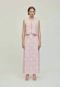 Vestido de crochê rosa com um colete curto e saia longa, apresentando recortes quadrados e uma textura suave. A roupa é complementada com sandálias de tiras.
