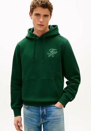 Jeune homme portant un sweat à capuche vert foncé avec poche avant et initiales brodées, associé à un jean bleu clair, debout devant un fond blanc.