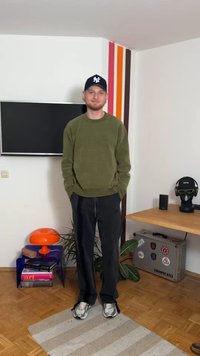 Olívazöld kötött pulóver, fekete nadrág, szürke sportcipők. Csíkos szőnyegen áll, mellette narancssárga lámpa, fekete fejhallgató és cserepes növény.