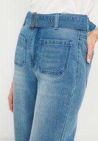 Γυναίκα που φοράει ανοιχτό μπλε τζιν παντελόνι ψηλής μέσης με φαρδιά ζώνη και τετράγωνη αγκράφα, με το χέρι στη μέση της, σε λευκό φόντο.