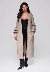 Langer beiger Strickcardigan mit offenem Frontbereich, gerippten Bündchen und zwei vorderen Taschen. Getragen über einem schwarzen Oberteil und dunklen Hosen mit spitzen schwarzen Stiefeln.