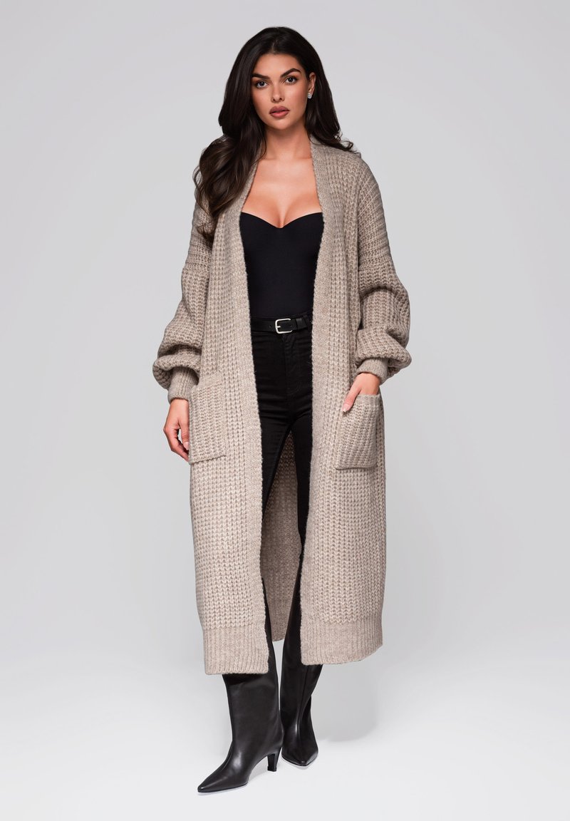 Langer beiger Strickcardigan mit offenem Frontbereich, gerippten Bündchen und zwei vorderen Taschen. Getragen über einem schwarzen Oberteil und dunklen Hosen mit spitzen schwarzen Stiefeln.