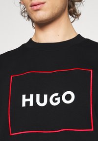 Sudadera negra con un gran logo blanco "HUGO" contorneado en costuras rojas. Corte regular, cuello de canalé y textura suave.