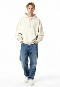 Felpa con cappuccio beige chiaro con tasca frontale e coulisse, abbinata a jeans di denim blu larghi e sneakers bianche. Design e vestibilità minimalisti.