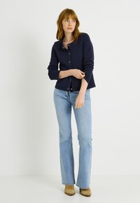 Cárdigan acolchado azul marino con escote redondo, cierre de botones y mangas largas. Combinado con jeans acampanados azul claro y botas beige hasta el tobillo.