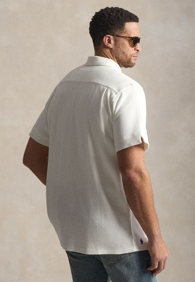 Λευκό πόλο με κοντά μανίκια από υφασμένο ύφασμα, με κλασικό γιακά, διακοσμημένο με μικρό κεντημένο λογότυπο κοντά στο τελείωμα.