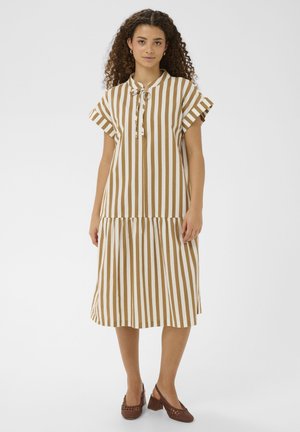 Kvinde med krøllet hår iført en beige og hvid midi-kjole med lodrette striber, korte ærmer og brune sko med hæl, stående.