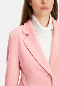 Helle rosa Cordjacke mit Revers, einem Ein-Knopf-Verschluss und gerippter Textur. Getragen über einem weißen Rollkragenpullover.