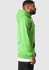 Felpa in cotone verde brillante, con tasca frontale, cappuccio con coulisse e polsini a costine. Indossata sopra una maglietta bianca, mostrata di profilo.