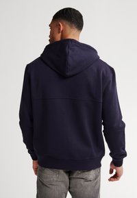 Sudadera azul marino con capucha, que presenta un ajuste ceñido y puños acanalados. Tejido de algodón grueso con una textura suave, detalles de diseño mínimos.