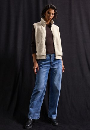 Gilet en polaire beige avec col montant, associé à un haut marron foncé et un jean bleu à jambes larges. Des bottes à talons noires complètent la tenue.