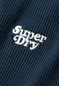 Tissu côtelé bleu marine foncé avec le texte brodé en blanc "Superdry", mettant en valeur une surface texturée avec un motif subtil.