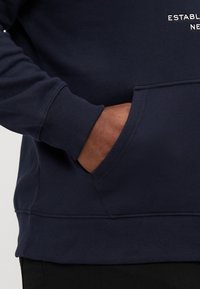 Sötétkék pulóver, puha pamut keverékből készült, bordázott mandzsettákkal és kenguruzsebbel. A mellkason lévő hímzett szöveg olvasható: "ALAPÍTVA."