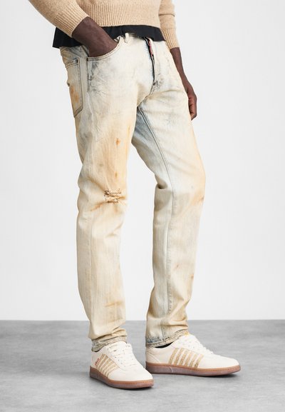 Jean en denim bleu clair avec des patchs déchirés et une décoloration, offrant un design classique à cinq poches et une fermeture éclair. Porté avec des baskets crème.