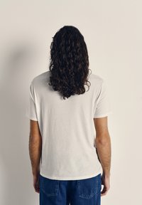 Bela majica s kratkimi rokavi, sproščen kroj, enobarvna tkanina. Prikazana od zadaj, s temi kodrastimi lasmi, ki padajo čez ramo.