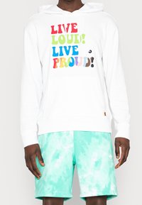 Person som bär en vit hoodie med multikolorigt text "LIVE LOUD! LIVE PROUD!" och ljus turkos batikmönstrade Adidas-shorts.