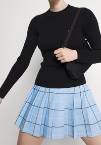 Mujer con una camiseta negra ajustada de manga larga y una falda ligera azul clara con líneas de cuadrícula negra, sosteniendo un bolso negro de hombro.